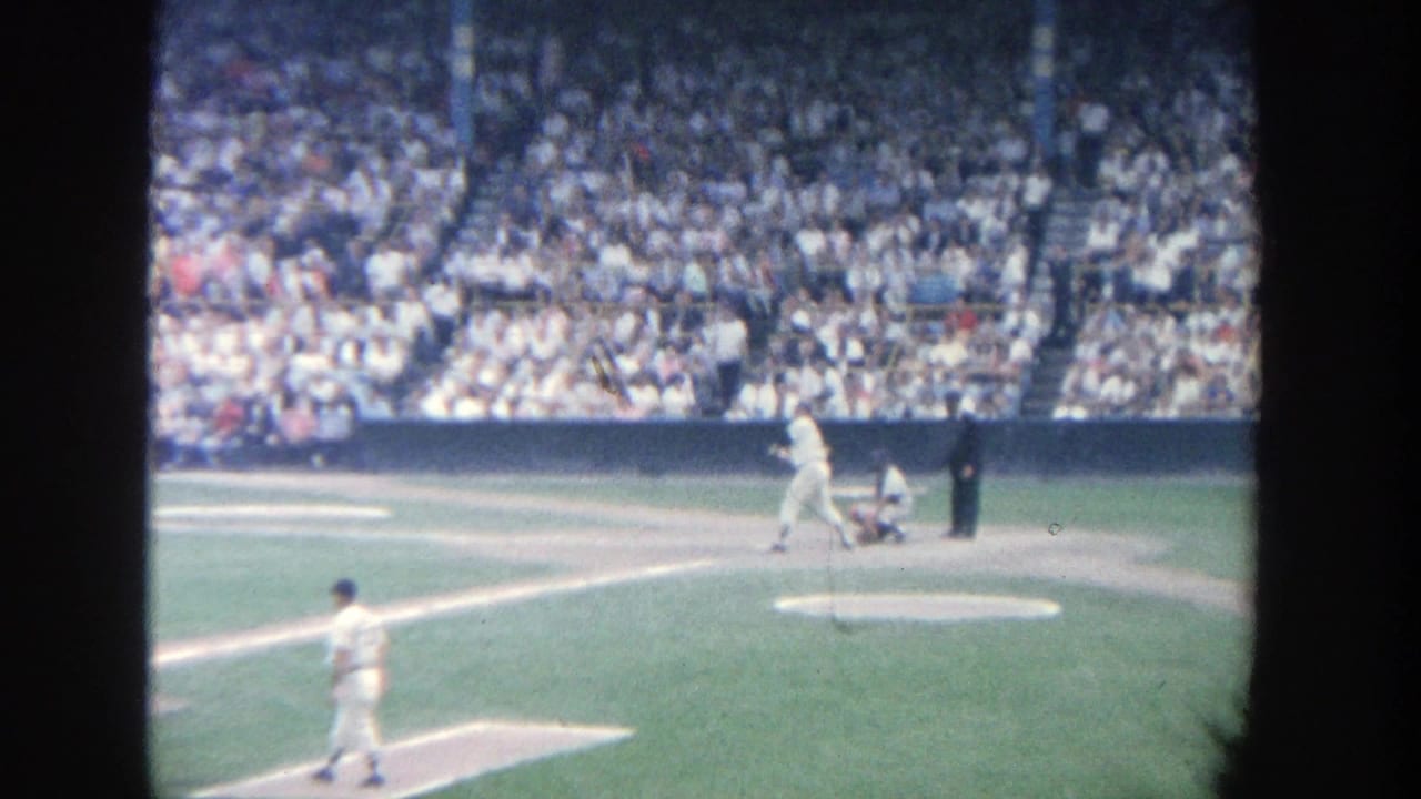 Batter swings near home plate as pitcher, catcher, umpire, and crowded stands are visible.
