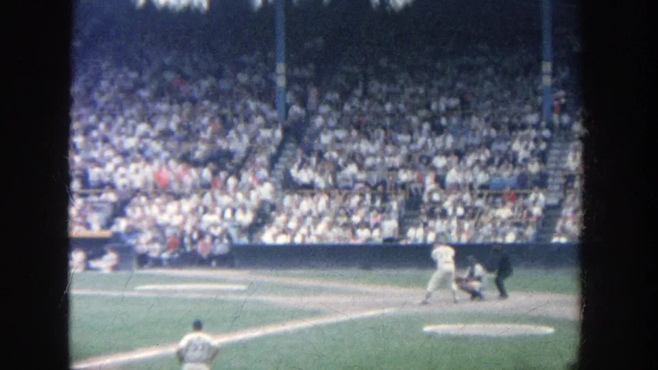 1964 Yankee Stadium frame with pitcher in the foreground and batter, catcher, umpire, and packed spectators across the field.