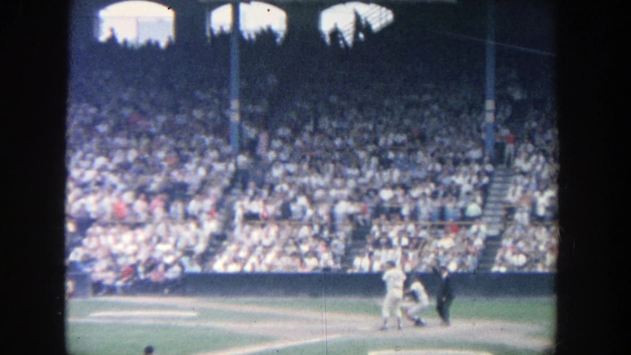 1964 Yankee Stadium home-movie frame showing a baseball play near home plate with dense spectator stands in the background.