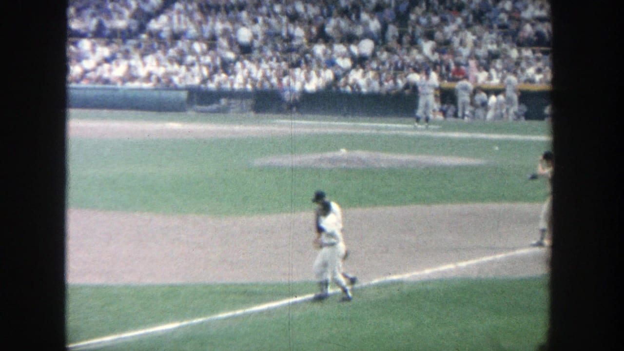 Baseball player in a white uniform walks along the baseline as players gather near the dugout.