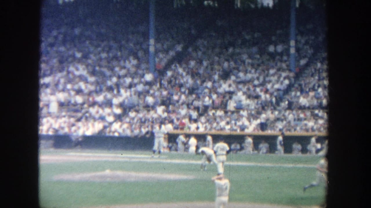1964 Yankee Stadium home-movie frame showing a pitcher preparing on the mound with players and packed stands in the background.