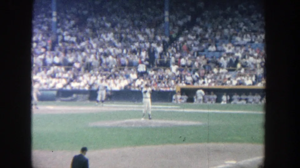 1964 Yankee Stadium 8mm frame with the pitcher centered on the mound, infield dirt below, and spectators filling the background.