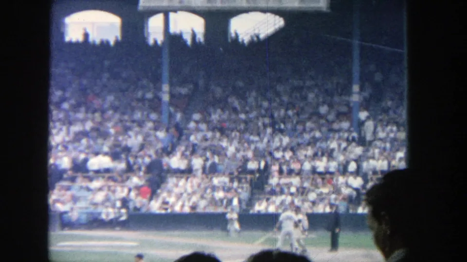 1964 Yankee Stadium home-movie frame showing the batter area, players near home plate, an umpire, and dense grandstand spectators.