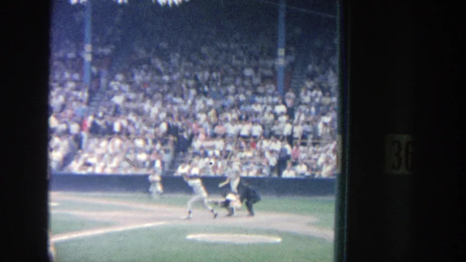 1964 8mm Yankee Stadium view of a batter swinging at home plate with catcher, umpire, pitcher mound, and packed grandstand visible.