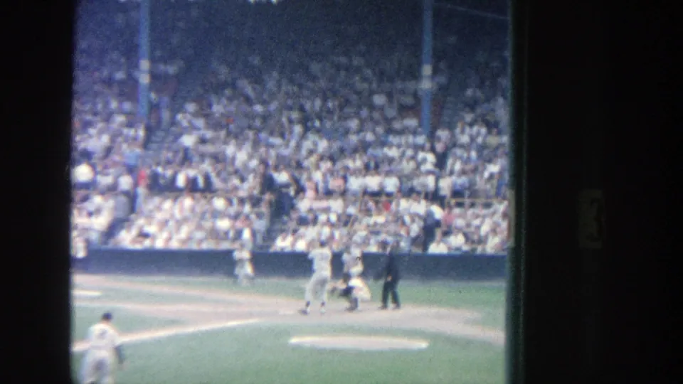 1964 Yankee Stadium home-movie frame showing a batter at home plate with catcher, umpire, fielders, and a packed crowd beyond.