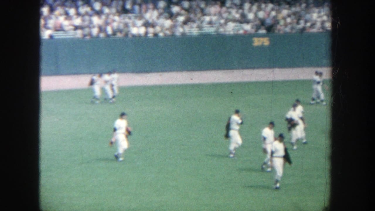 Baseball players in white uniforms walking across a green field with spectators behind them.