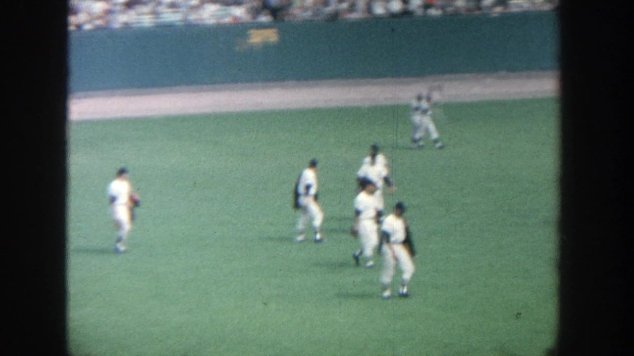 Several baseball players stand and walk on the outfield grass near a green wall.