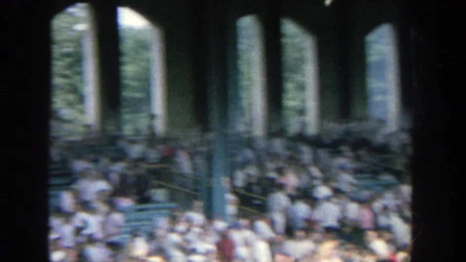 A blurred 8mm view inside Yankee Stadium shows spectators, railings, seating rows, and vertical structural columns from a spectator vantage point.