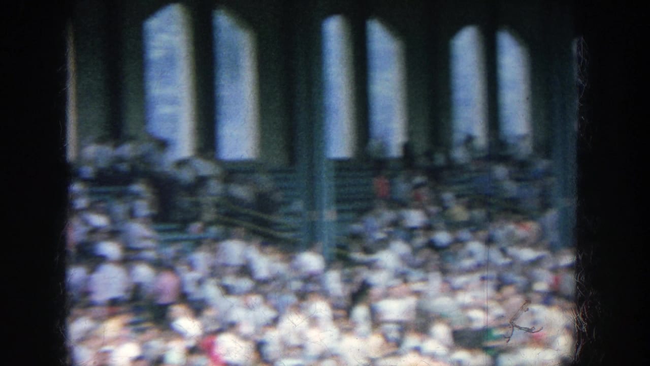 1964 Yankee Stadium home-movie frame showing crowded bleachers, arched openings, and dark foreground window or pillar shapes.