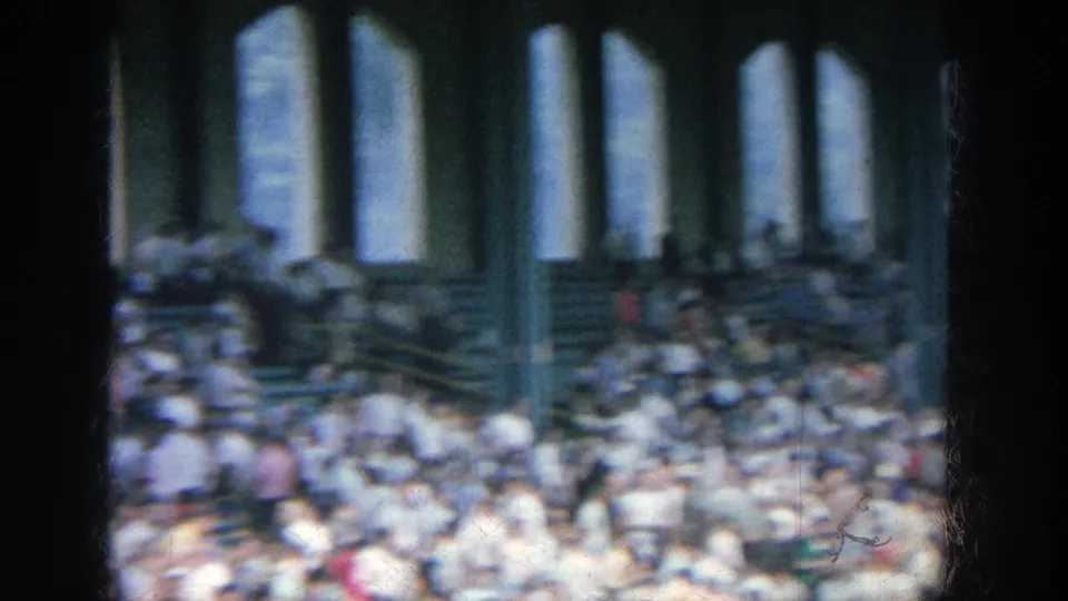 1964 Yankee Stadium home-movie frame showing crowded bleachers, arched openings, and dark foreground window or pillar shapes.
