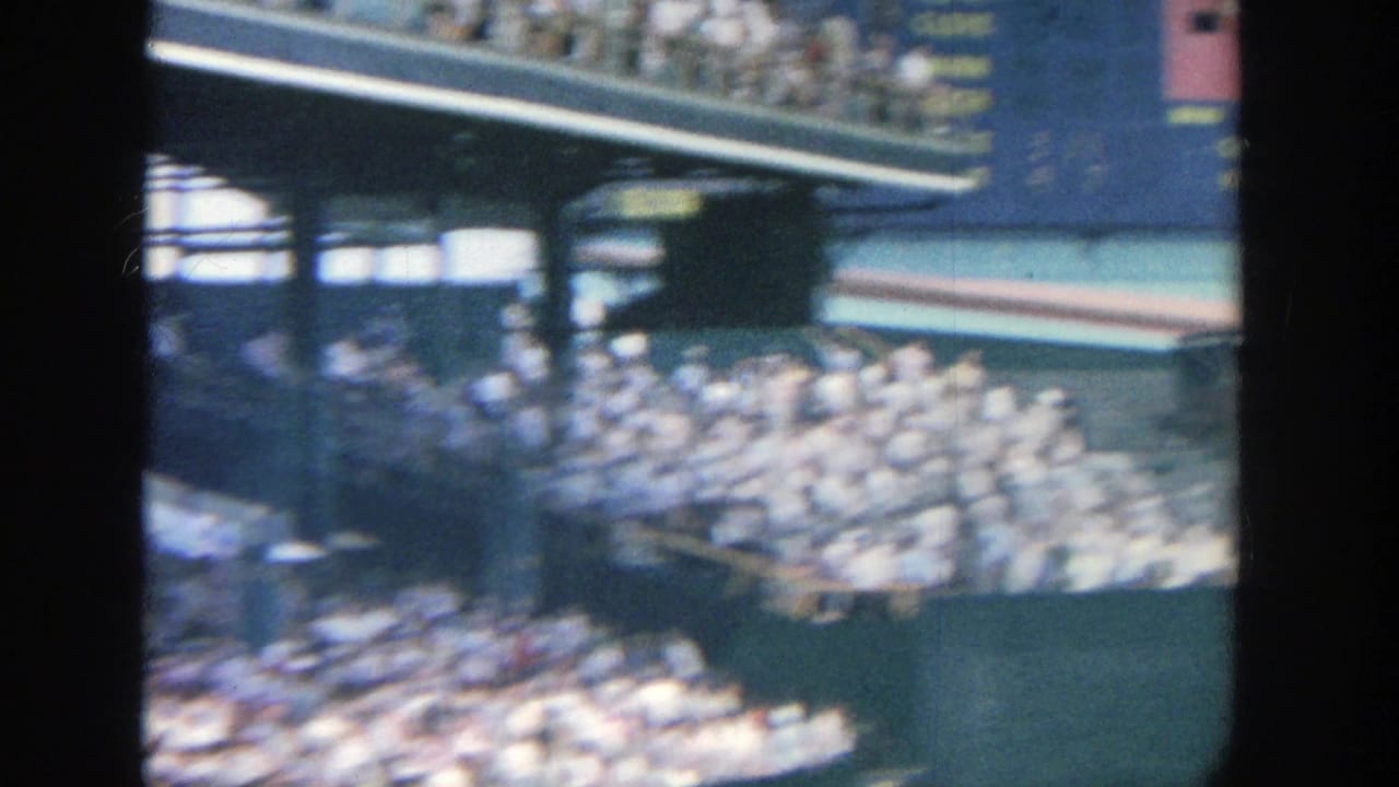 1964 Yankee Stadium frame with spectators filling the stands beneath a large scoreboard or sign structure during the pan.