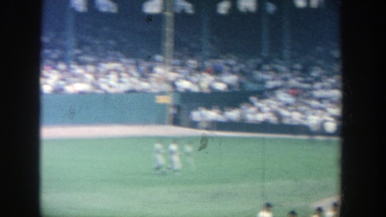 Baseball field seen from the stands with players in white uniforms and a packed crowd beyond.