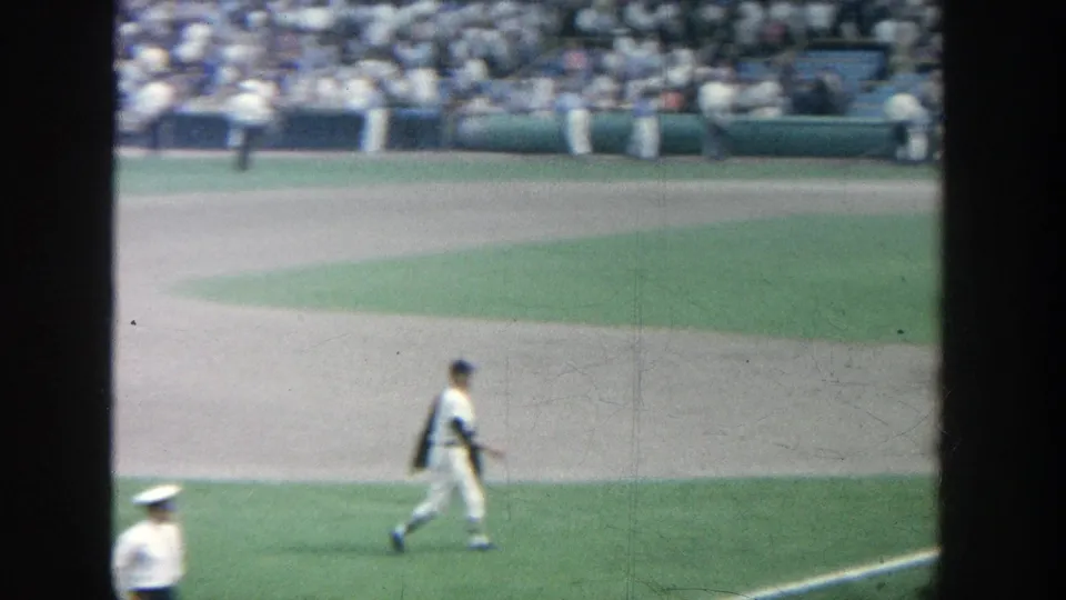 1964 Yankee Stadium home-movie frame showing a player walking near the infield, with a uniformed attendant partly visible below.
