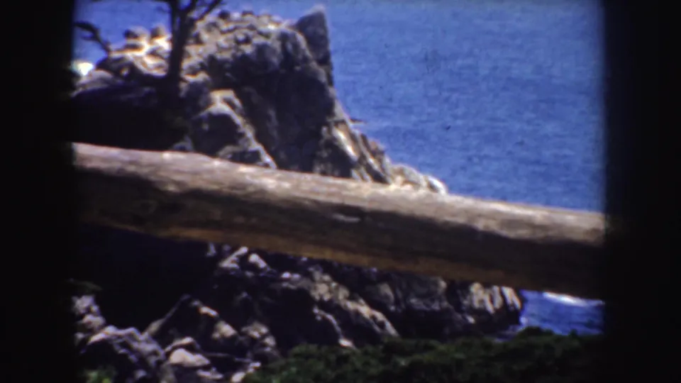 1952 California coastal frame showing the rocky point, deep blue ocean, railing, and a partial view of the cypress.