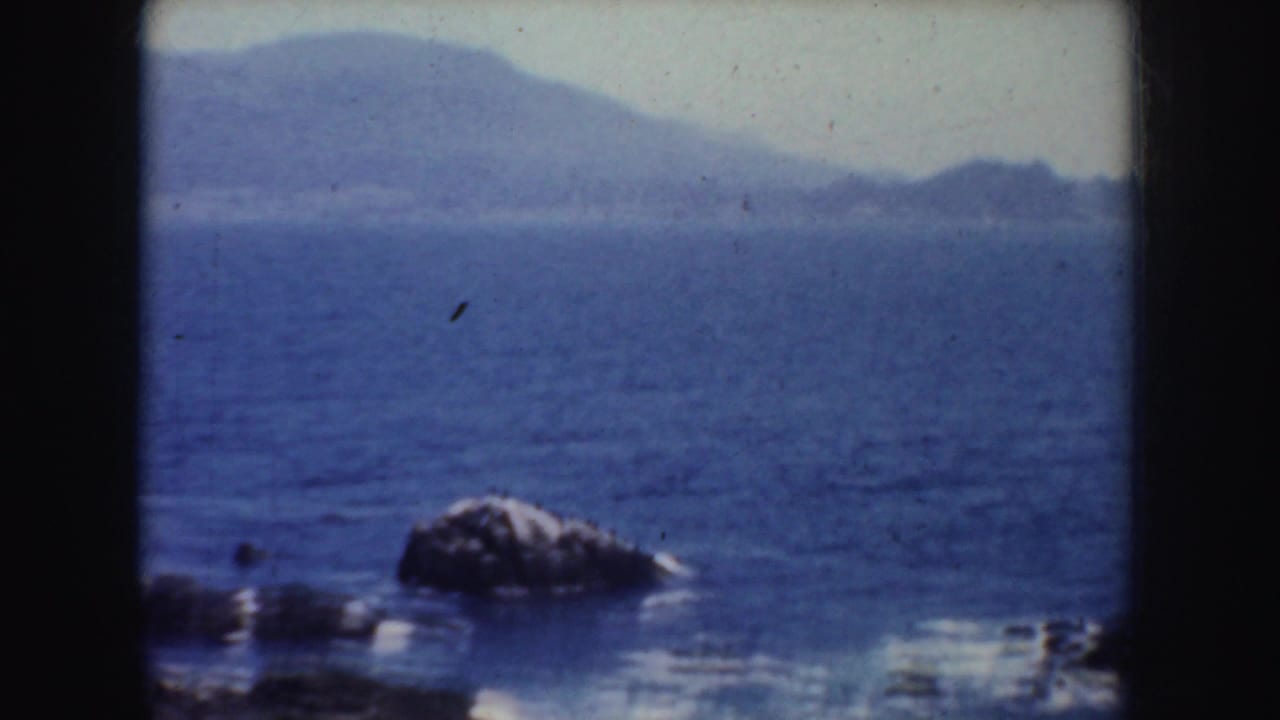 1952 California 8mm frame showing blue water, a rocky shoreline detail, and hazy hills beyond the coast.