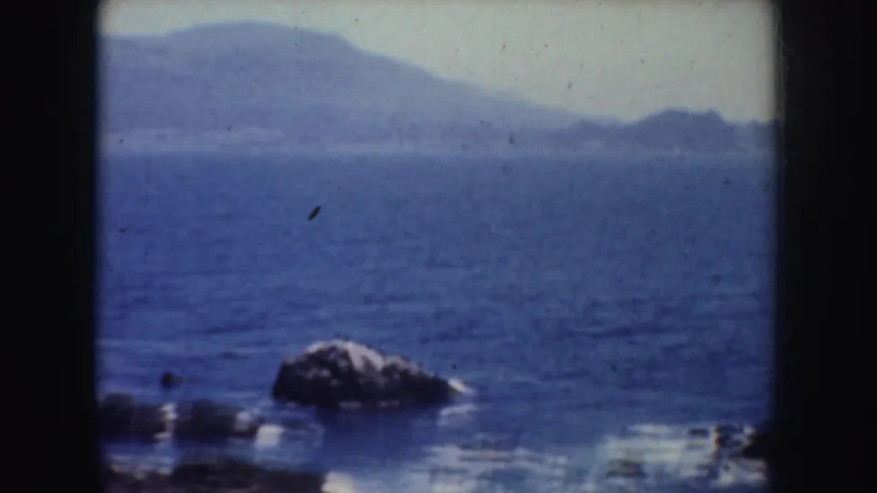 1952 California 8mm frame showing blue water, a rocky shoreline detail, and hazy hills beyond the coast.