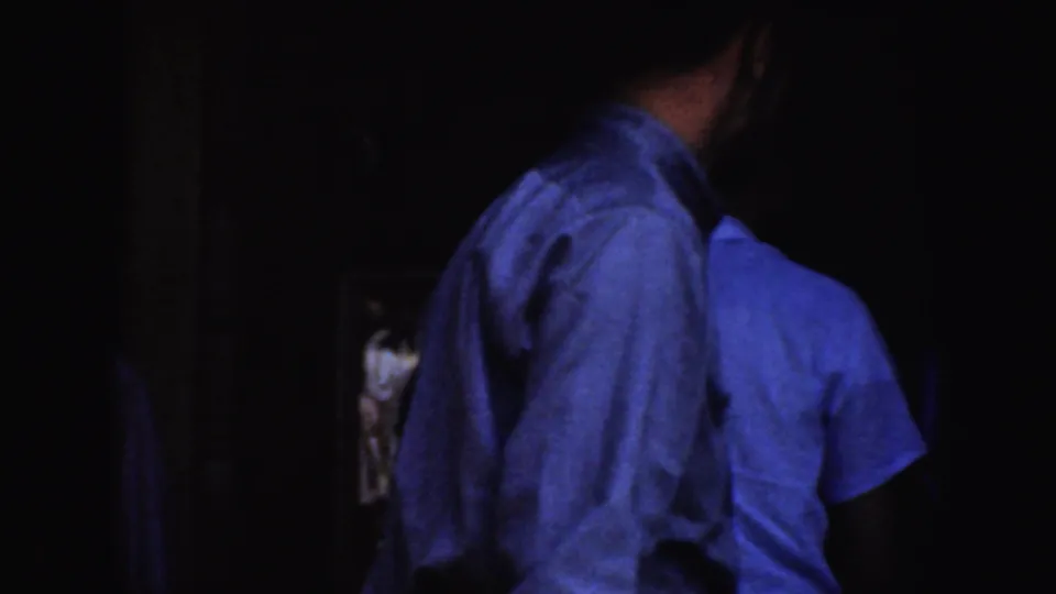 Dim 1952 California home-movie frame with adult men in light shirts near the saloon doorway and partial sign area.