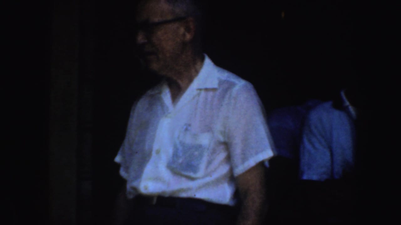 Older man in glasses stands in shadow near other visitors.