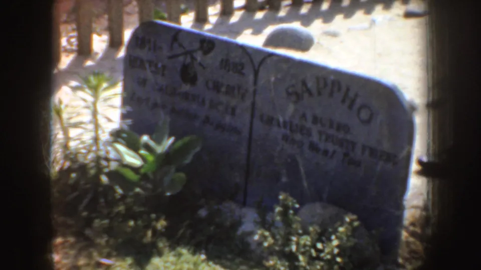 1952 California frame with the Sappho marker and another grey tombstone partly framed by a wooden fence.