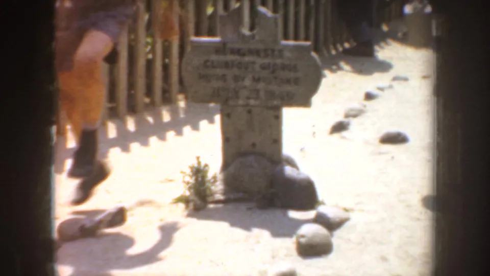 1952 California 8mm frame showing one grey marker near a fence, rocks, sandy ground, and a partial passerby at left.
