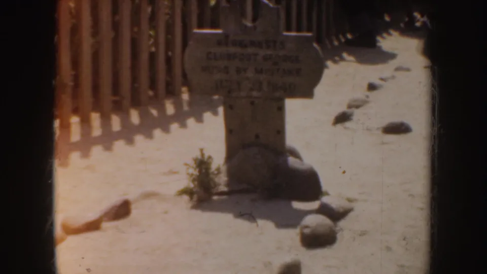 1952 California frame showing a small grey tombstone by a wooden fence with rocks, plant growth, and strong sunlight.
