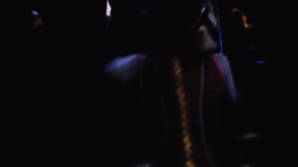 A dim 8mm frame shows a partial performer or handler shape with colored trim and a small bright highlight in shadow.