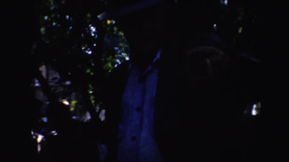 Low-light 1952 California home-movie frame with a jacketed figure, hat shape, foliage, and scattered highlights.