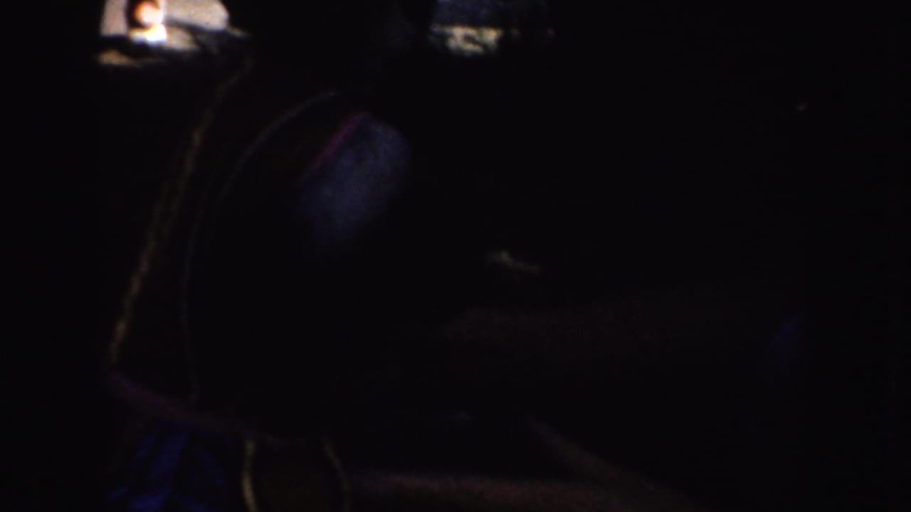 In a dark 1952 California home-movie frame, a seated woman appears to handle blue fabric while wearing a white sleeveless shirt.