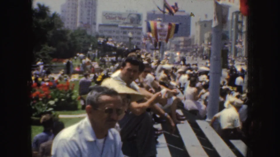 1952 California crowd scene with men seated near steps, flower beds, flags, bunting, and a city street filled with spectators.