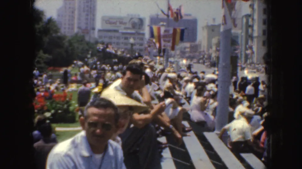1952 California home-movie frame showing seated spectators, a dense street crowd, flags, bunting, flowers, and urban buildings.