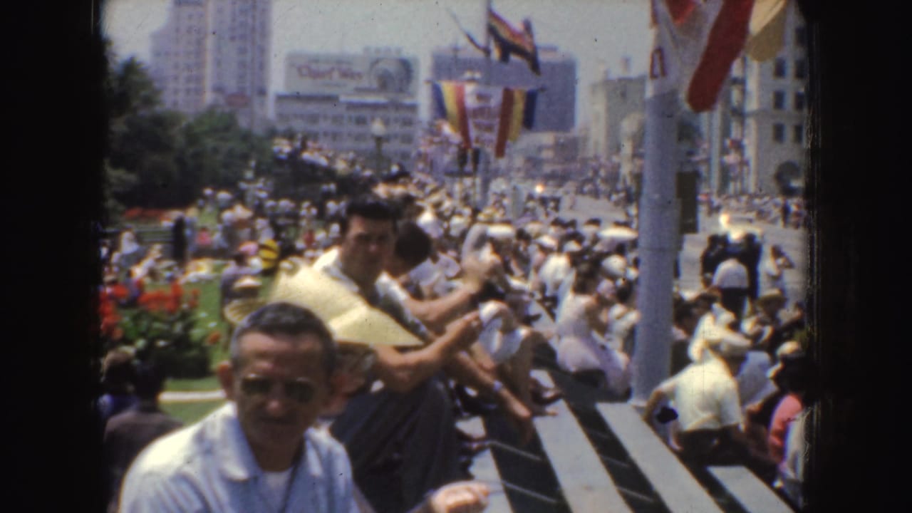 1952 California home-movie frame showing spectators on bleachers along a city street with colorful flags and buildings in the background.