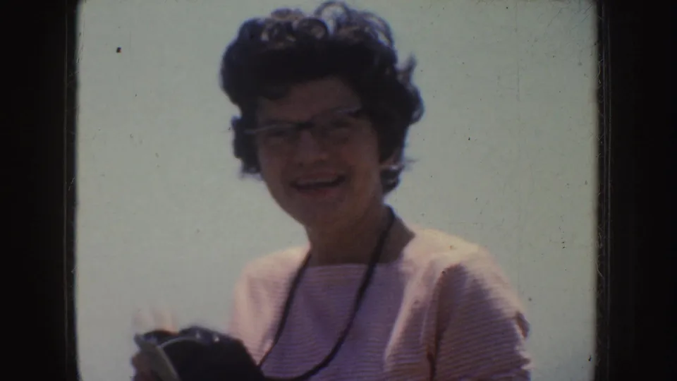 A smiling woman in glasses and a striped top holds a camera in a sunlit 1952 California home-movie frame.