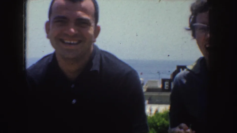 A smiling man in a dark polo faces the camera, with another adult partly visible beside him and a bright California horizon behind.