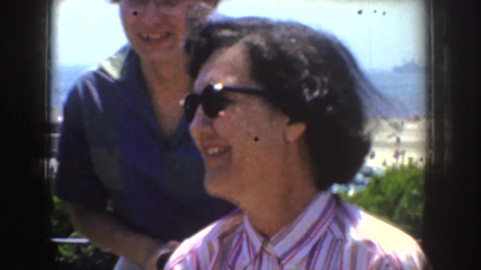Woman in dark sunglasses smiles in bright sun with another adult nearby and the California coast behind them.