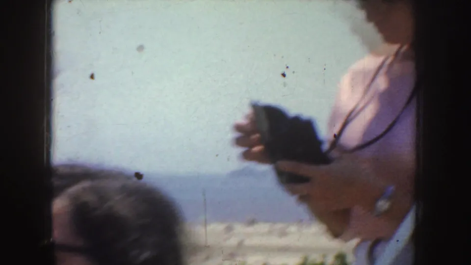 Cropped 1952 California frame showing an adult holding a small camera against a bright ocean backdrop.