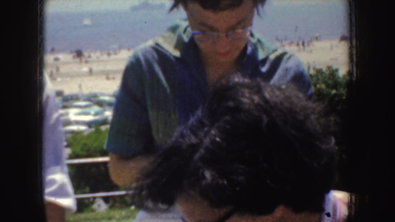 1952 California home-movie frame of a woman in glasses seated near bleachers, with beach activity and parked cars behind her.