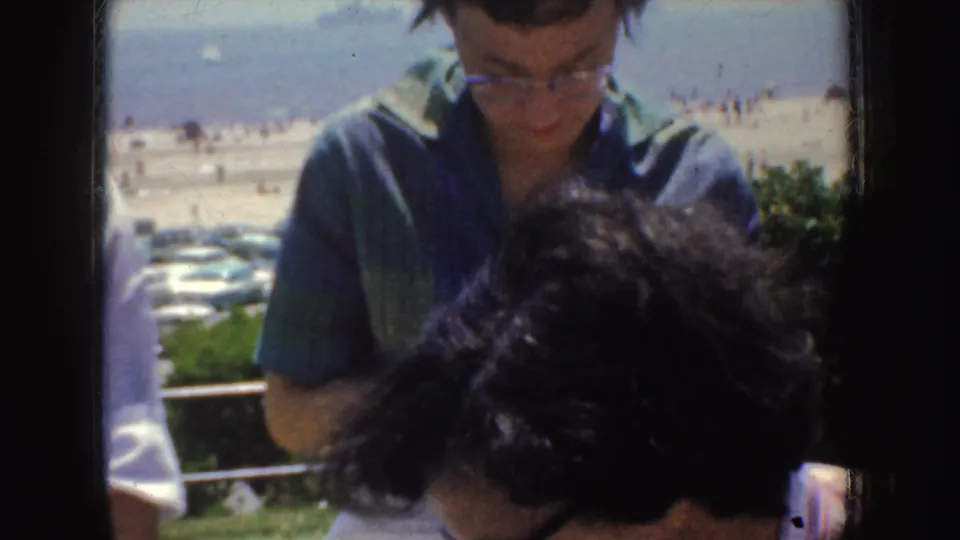 1952 California home-movie frame of a woman in glasses seated near bleachers, with beach activity and parked cars behind her.