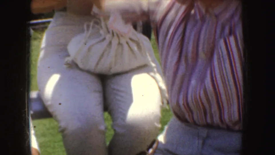 Close 1952 California frame of seated spectators on grass, with a striped blouse, light trousers, and a gathered purse visible.