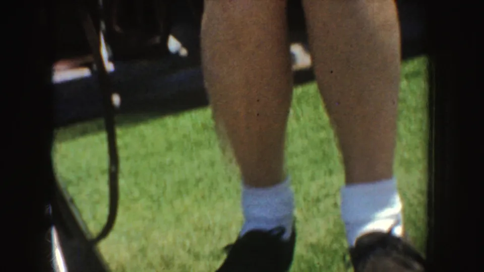 1952 California home-movie detail of seated legs with white socks and dark shoes near a bleacher on grass.