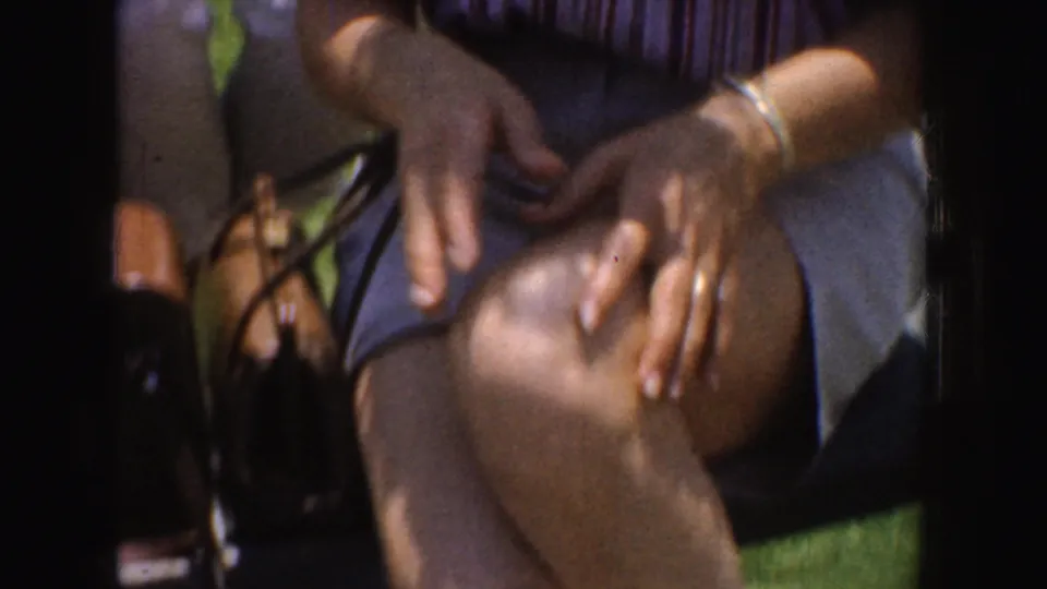 1952 California frame of a seated woman in a striped shirt with crossed legs, bracelets, purse strap, and hands resting in her lap.