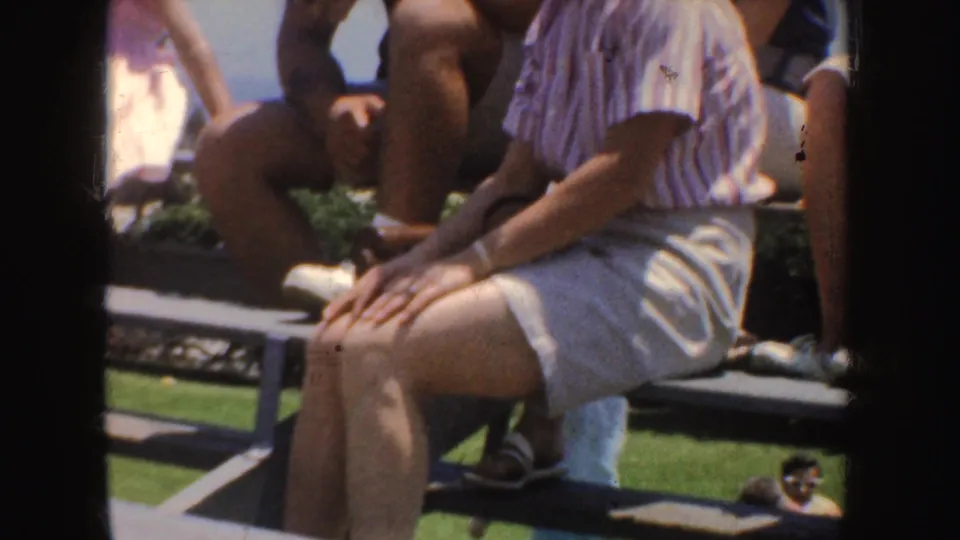 1952 California home-movie frame showing adults on a bench, with cropped figures, summer clothing, and grass visible.