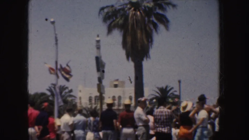 1952 California beach crowd in summer clothing gathered near palm trees, flags, and low buildings facing the ocean.