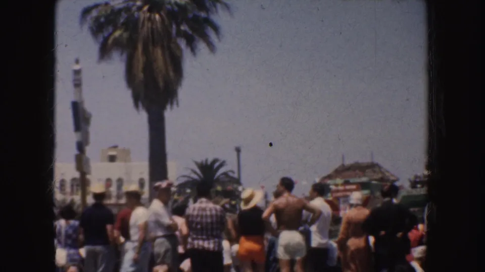 Crowd on a 1952 California beach faces the water, with palm trees, hats, swimwear, and a small beach structure visible.