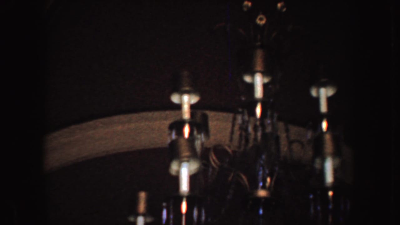 1952 California 8mm frame showing a dim interior with part of a hanging chandelier, pale bulbs, and dark surrounding surfaces.