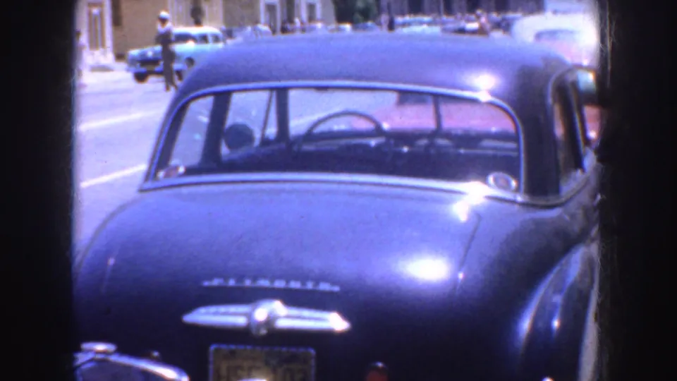1952 California street view with the rear of a blue sedan, sunlit pavement, and distant parked cars.