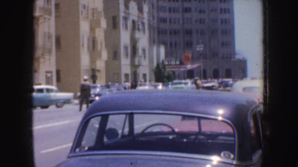 1952 California street scene with a blue sedan, tan apartment buildings, pedestrians, and parked cars.