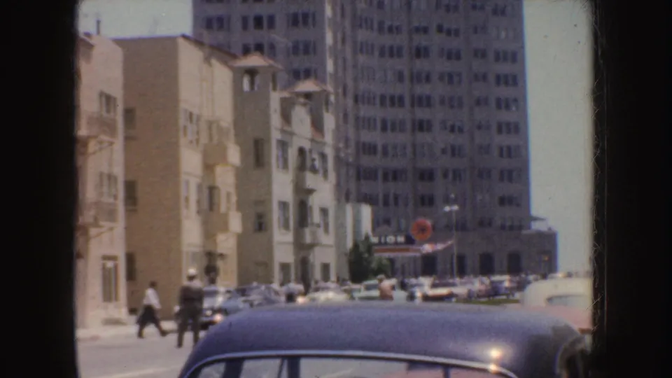 1952 California urban view showing a blue car in the foreground, pedestrians, tan buildings, and a taller background facade.