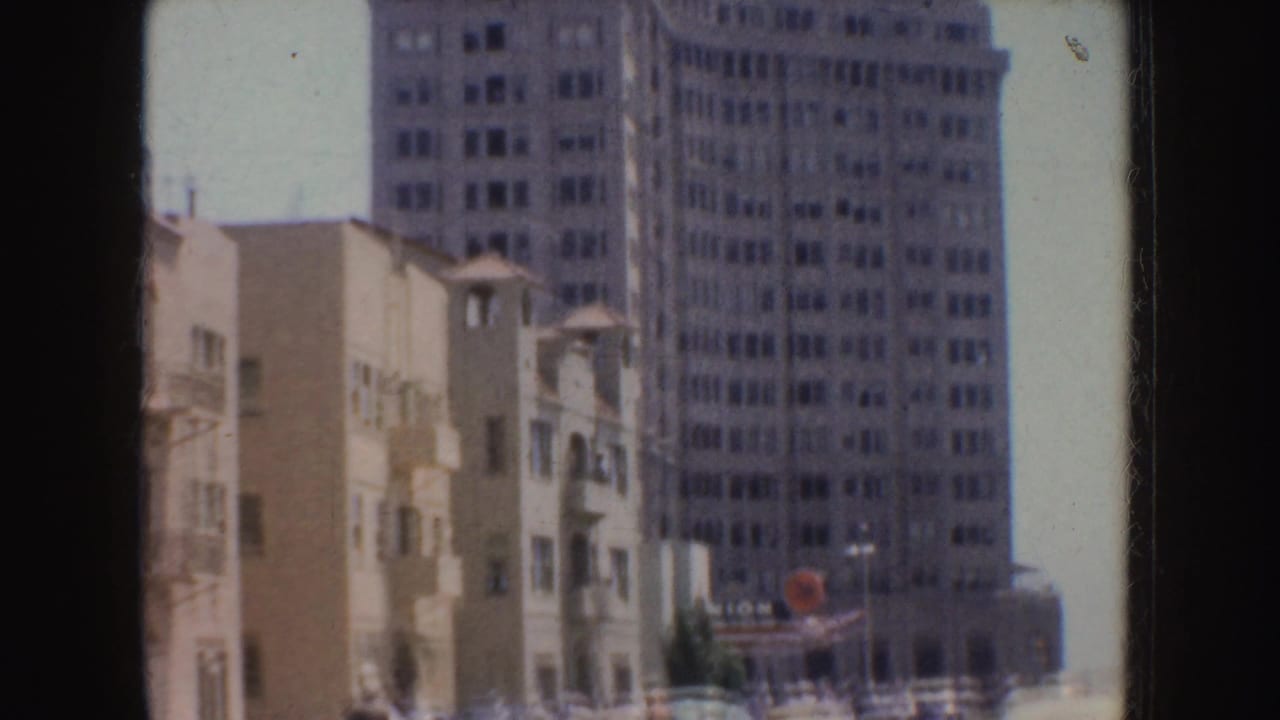 1952 California 8mm frame showing a tall multi-window building behind smaller pale urban buildings in bright daylight.