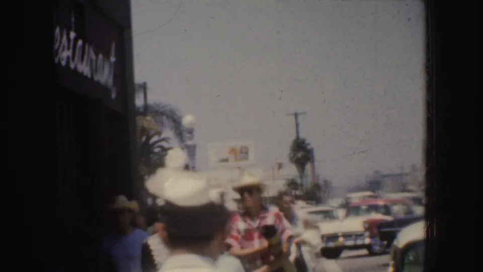 1952 California sidewalk crowd with men in hats, a partial restaurant sign, palm-like street planting, and classic cars in traffic.