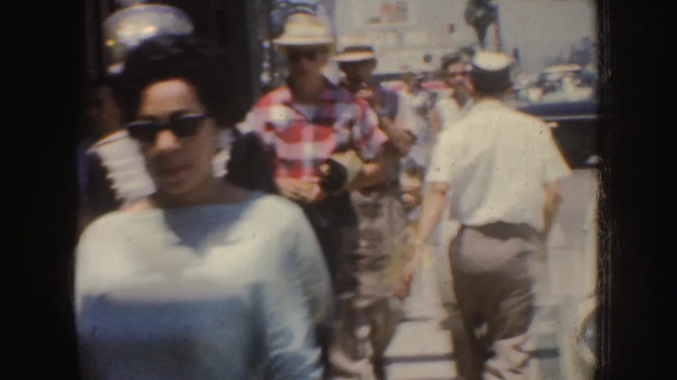 A woman in sunglasses walks past the camera as 1950s California pedestrians continue along a crowded downtown sidewalk.